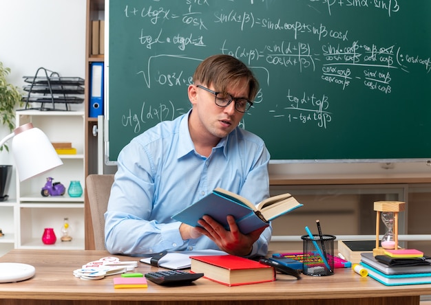 Foto grátis jovem professor usando óculos, lendo um livro, preparando a aula, parecendo confiante sentado na mesa da escola com livros e notas na frente do quadro-negro na sala de aula