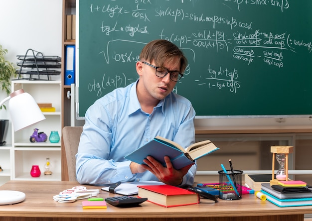Jovem professor usando óculos, lendo um livro, preparando a aula, parecendo confiante sentado na mesa da escola com livros e notas na frente do quadro-negro na sala de aula