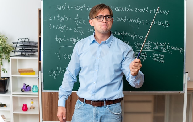 Jovem professor zangado usando óculos com um ponteiro explicando a lição em pé perto do quadro-negro com fórmulas matemáticas na sala de aula