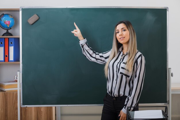 jovem professora em pé perto do quadro-negro na sala de aula explicando a lição apontando para o quadro-negro com o dedo indicador parecendo confiante