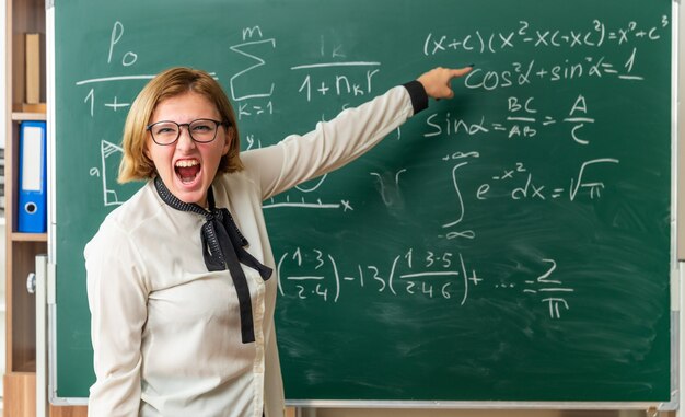 Jovem professora furiosa usando óculos em pé na frente do quadro-negro aponta para o quadro-negro na sala de aula