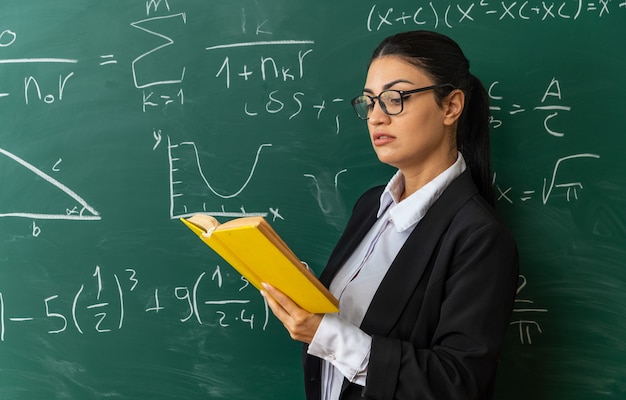 Jovem professora impressionada usando óculos em pé na frente da lousa lendo um livro na sala de aula