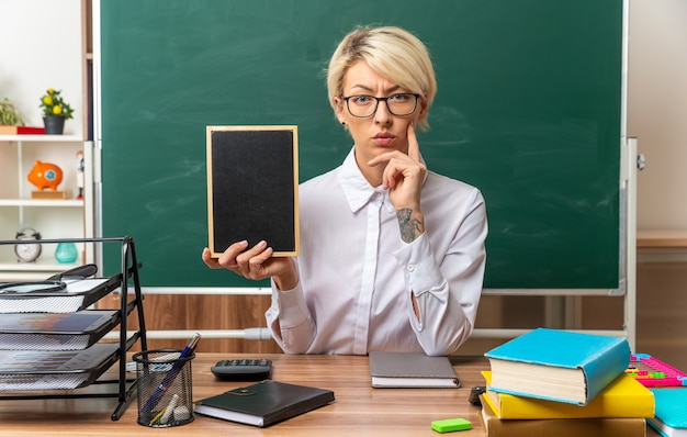 jovem professora loira pensativa usando óculos, sentada na mesa com o material escolar na sala de aula, mostrando uma minilousa com a mão no queixo, olhando para a frente