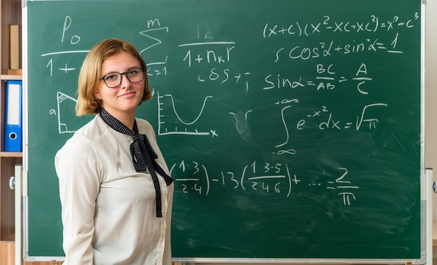 Jovem professora satisfeita usando óculos, parada na frente do quadro-negro na sala de aula