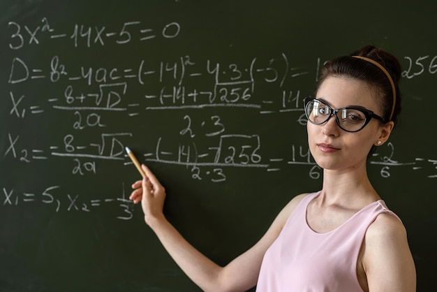 Jovem professora séria explica fórmulas matemáticas na universidade ao lado do quadro-negro Foto Premium