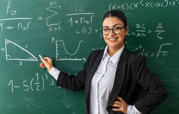 Jovem professora sorridente usando óculos, parada na frente do quadro-negro, segurando encalhado para a prancha colocando a mão no quadril na sala de aula