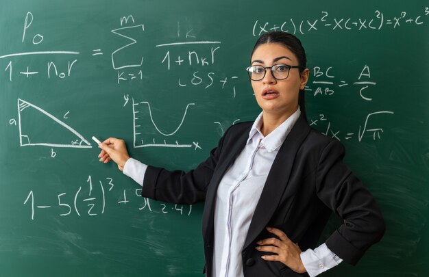 Jovem professora surpresa usando óculos em pé na frente do quadro-negro segurando encalhado para a prancha colocando a mão no quadril na sala de aula