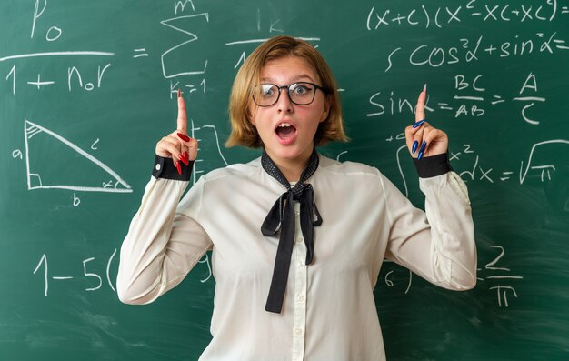 Jovem professora surpresa usando óculos, parada na frente do quadro negro, apontando para cima na sala de aula