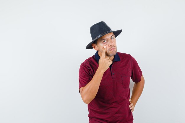 Jovem puxando para baixo a pálpebra inferior com o dedo na camiseta, chapéu e olhando sarcástico, vista frontal.