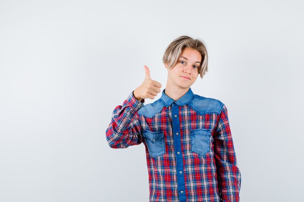 Jovem rapaz adolescente aparecendo o polegar em uma camisa e olhando alegre, vista frontal.