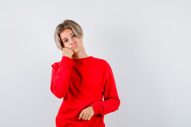 Jovem rapaz adolescente apoiando a bochecha no punho no suéter vermelho e parecendo triste. vista frontal.