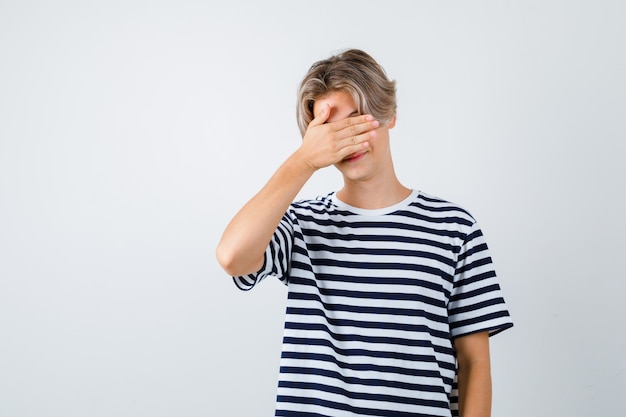 Jovem rapaz adolescente com a mão no rosto em uma camiseta listrada e parecendo envergonhado. vista frontal.