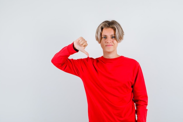 Foto grátis jovem rapaz adolescente com suéter vermelho, mostrando o polegar para baixo e parecendo descontente, vista frontal.