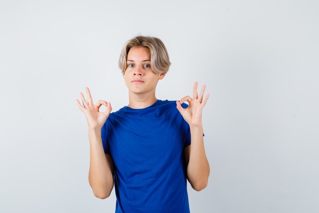 Jovem rapaz adolescente em t-shirt azul, mostrando o gesto de ok e olhando espantado, vista frontal.