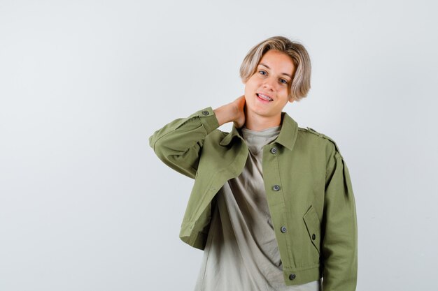 Jovem rapaz adolescente mantendo a mão atrás do pescoço em t-shirt, jaqueta e parecendo jovial, vista frontal.
