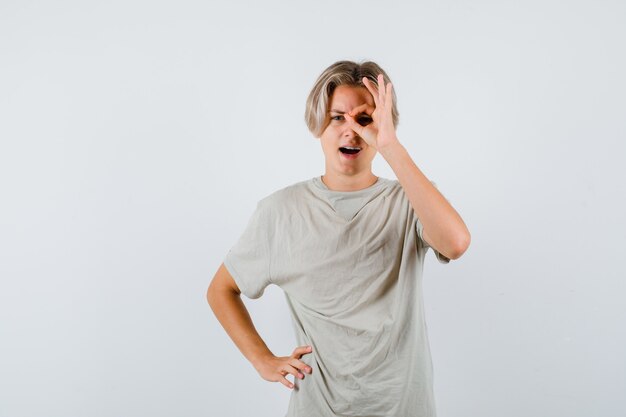 Jovem rapaz adolescente mostrando sinal ok no olho em t-shirt e olhando curioso, vista frontal.