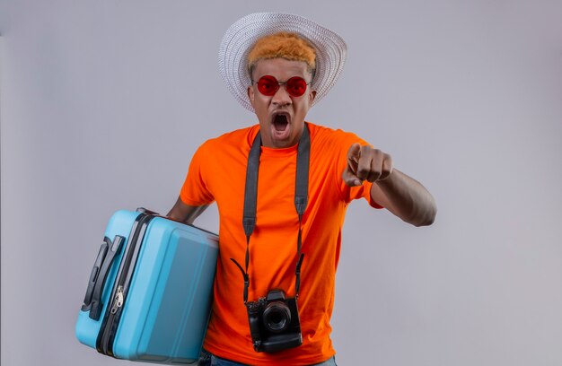 Jovem rapaz bonito com chapéu de verão e camiseta laranja segurando uma mala de viagem, apontando para a câmera com um dedo gritando com cara de raiva