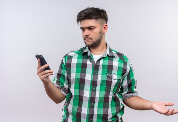 Jovem rapaz bonito de camisa quadriculada e surpreso com o telefone em pé sobre uma parede branca