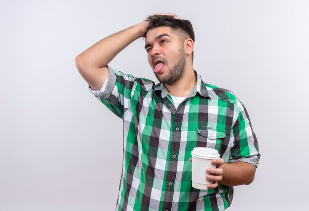 Jovem rapaz bonito de camisa quadriculada tentando se lembrar de segurar uma xícara de café de plástico sobre uma parede branca