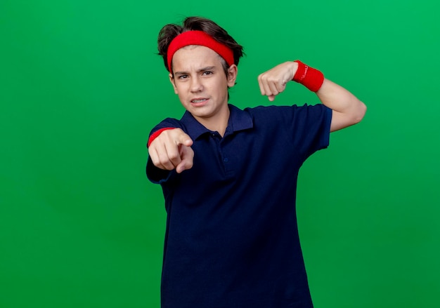 Foto grátis jovem rapaz bonito desportivo carrancudo usando bandana e pulseiras com aparelho dentário, fazendo um gesto forte, olhando e apontando para a frente, isolado na parede verde com espaço de cópia
