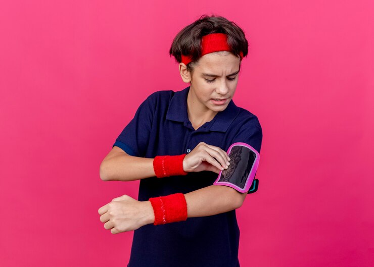 Foto grátis jovem rapaz bonito e carrancudo, desportivo, usando fita para a cabeça, pulseiras e uma braçadeira de telefone com aparelho dentário tocando a braçadeira isolada na parede carmesim com espaço de cópia