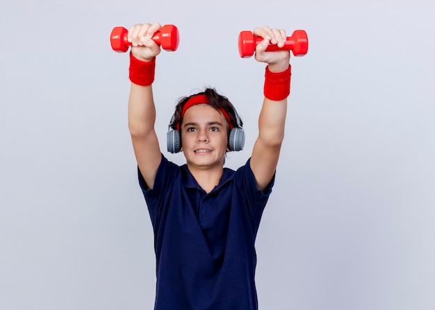 Foto grátis jovem rapaz bonito e desportivo concentrado usando bandana e pulseiras e fones de ouvido com aparelho dentário levantando halteres olhando diretamente isolado no fundo branco com espaço de cópia