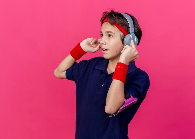 Foto grátis jovem rapaz bonito e desportivo impressionado usando bandana, pulseiras e fones de ouvido, braçadeira de telefone com aparelho dentário, olhando direto, tocando fones de ouvido isolados em um fundo carmesim com espaço de cópia