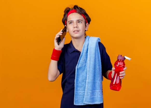 Jovem rapaz bonito e desportivo usando bandana e pulseiras com aparelho dentário e toalha no ombro segurando uma garrafa de água falando no telefone isolado na parede laranja com espaço de cópia