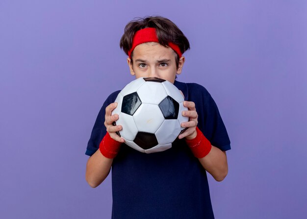 Jovem rapaz bonito e esportivo impressionado usando bandana e pulseiras com aparelho dentário segurando uma bola de futebol por trás, isolada na parede roxa com espaço de cópia