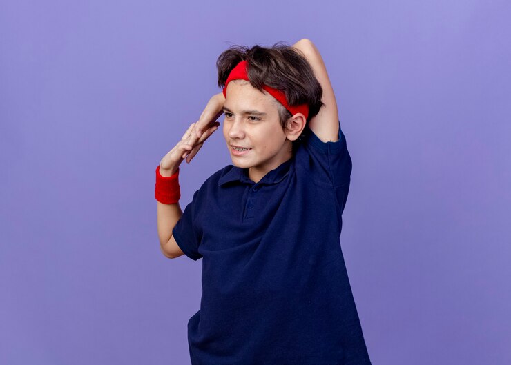 Foto grátis jovem rapaz bonito e esportivo satisfeito usando fita para a cabeça e pulseiras com aparelho dentário, olhando para o lado, mantendo as mãos juntas atrás do pescoço, exercitando-se isolado no fundo roxo com espaço de cópia