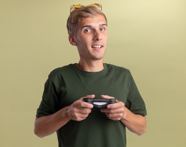 Jovem rapaz bonito satisfeito, vestindo uma camisa verde com óculos na cabeça, jogando no joystick do controlador de jogo isolado na parede verde oliva