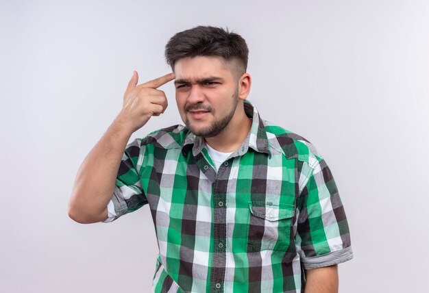 Jovem rapaz bonito vestindo camisa xadrez, olhando ofensivamente, além de pedir para pensar em pé sobre uma parede branca