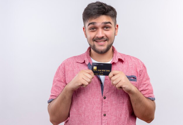 Jovem rapaz bonito vestindo uma camisa pólo rosa e olhando feliz segurando um cartão de crédito em pé sobre uma parede branca