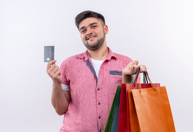 Jovem rapaz bonito vestindo uma camisa pólo rosa feliz procurando fazer compras com um cartão de crédito em pé sobre uma parede branca