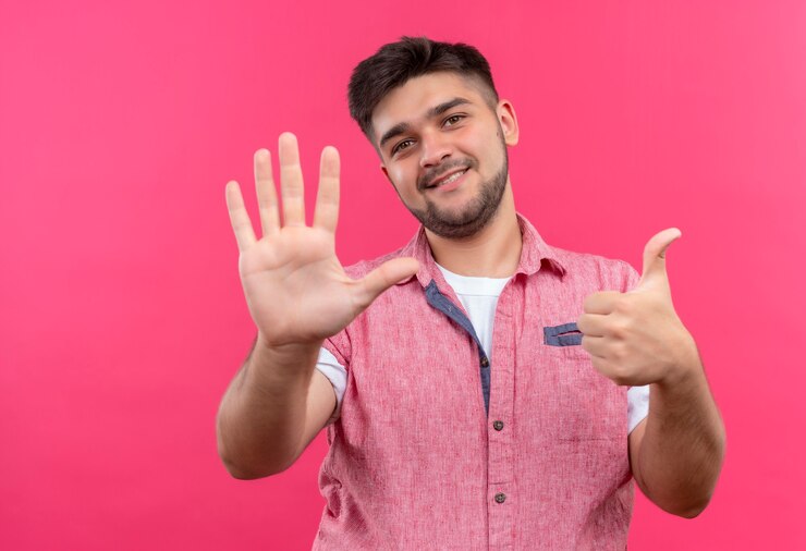 Foto grátis jovem rapaz bonito vestindo uma camisa pólo rosa mostrando cinco e um em pé sobre uma parede rosa