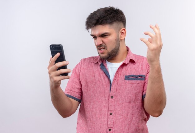 Jovem rapaz bonito vestindo uma camisa pólo rosa olhando para o telefone com raiva em pé sobre uma parede branca