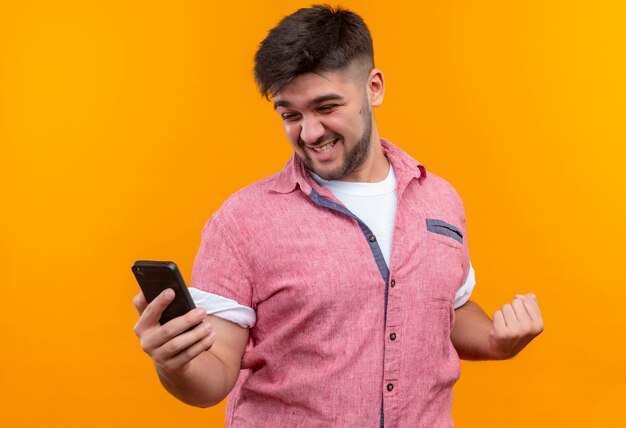 Jovem rapaz bonito vestindo uma camisa pólo rosa olhando para o telefone e se alegrando de pé sobre a parede laranja