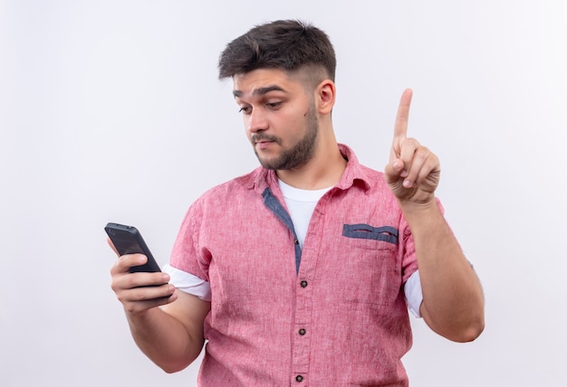 Jovem rapaz bonito vestindo uma camisa pólo rosa olhando para o telefone pensativamente levantando o dedo indicador em pé sobre a parede branca