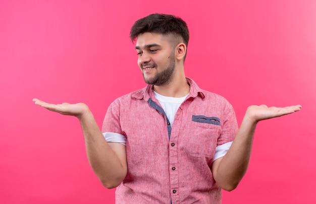 Jovem rapaz bonito vestindo uma camisa pólo rosa segurando algo nas mãos esquerda e direita, olhando para a direita em pé sobre a parede rosa