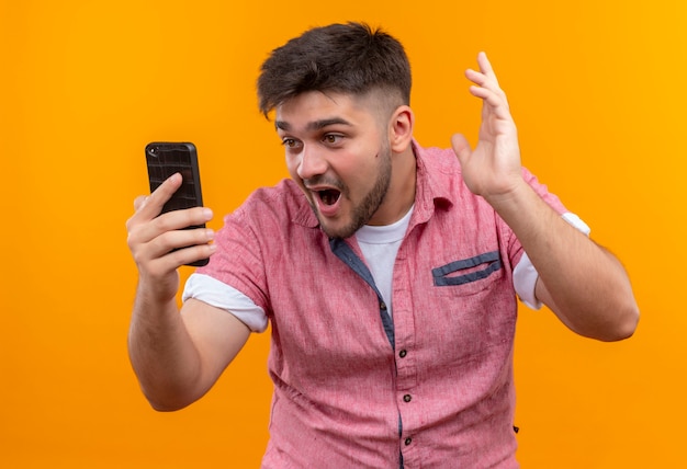 Foto grátis jovem rapaz bonito vestindo uma camisa pólo rosa surpreso olhando para o telefone em pé sobre a parede laranja