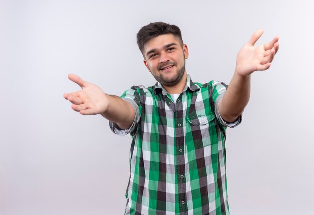 Jovem rapaz bonito vestindo uma camisa xadrez e olhando feliz abrindo os braços em cima da parede branca