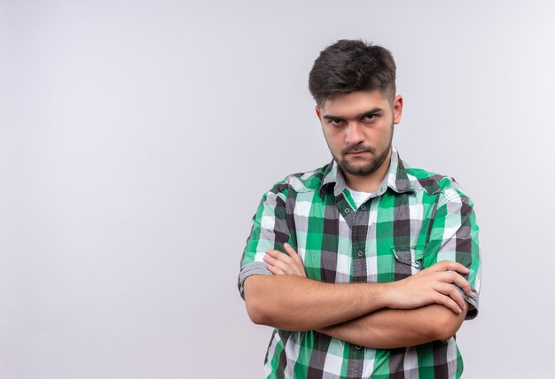 Jovem rapaz bonito vestindo uma camisa xadrez e parecendo ofendido em pé sobre uma parede branca