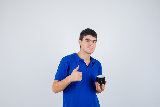 Jovem rapaz de t-shirt azul, segurando o copo, aparecendo o polegar e olhando feliz, vista frontal.