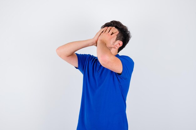 Jovem rapaz em t-shirt azul cobrindo parte do rosto com a mão e parecendo irritado, vista frontal.