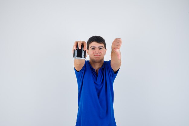 Jovem rapaz segurando a xícara, mostrando o polegar para baixo em uma camiseta azul e parecendo confiante, vista frontal.