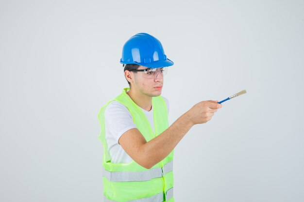 Jovem rapaz segurando o pincel, tentando dá-lo a alguém com uniforme de construção e olhando com foco.