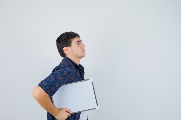 Jovem rapaz segurando whiteboard em t-shirt branca, camisa floral e olhando sério.