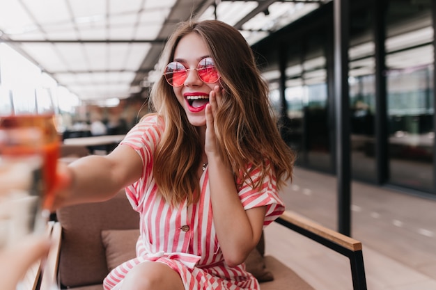 Jovem refinada em óculos de sol, comemorando algo no café. Foto interna de uma linda garota sorridente usa um vestido listrado de verão.