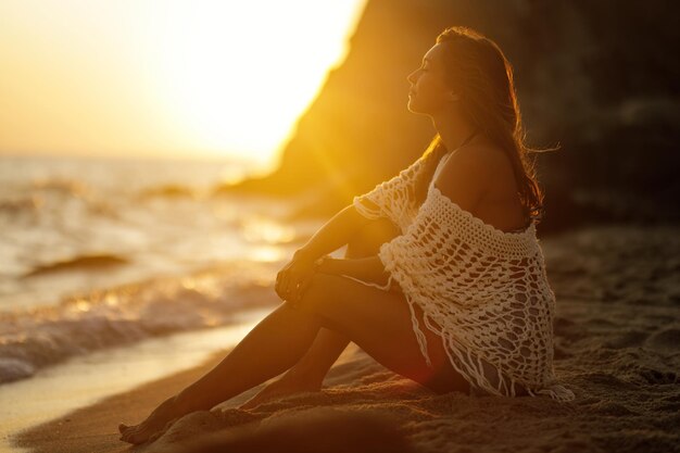 Jovem relaxada sentada na areia e curtindo o pôr do sol com os olhos fechados