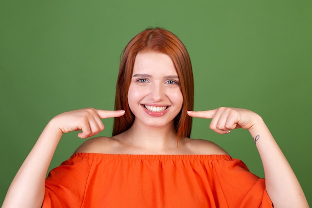 Jovem ruiva com blusa laranja casual na parede verde apontar dedos nos dentes brancos sorriso perfeito